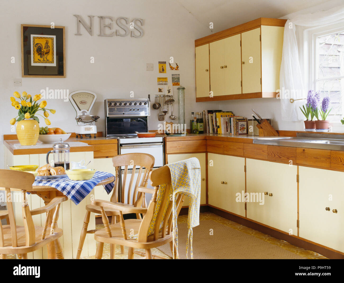 Pine chairs and table in country kitchen with metal letters on wall above small gas oven and traditional weighing scales Stock Photo