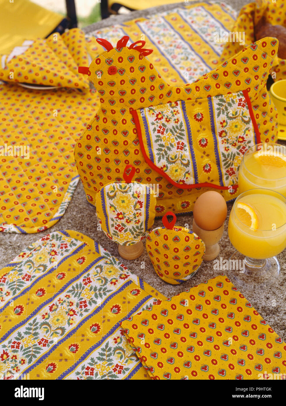 Close-up of yellow Provencal fabric egg-cosies on table beside hen-shaped tea-cosy and glasses of orange juice Stock Photo