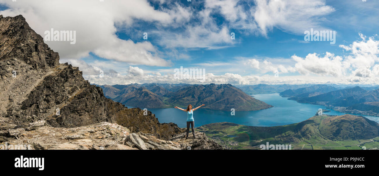 Female hiker stands on rocks in The Remarkables, overlooking Lake Wakatipu, mountains and Queenstown, Queenstown, Otago Stock Photo