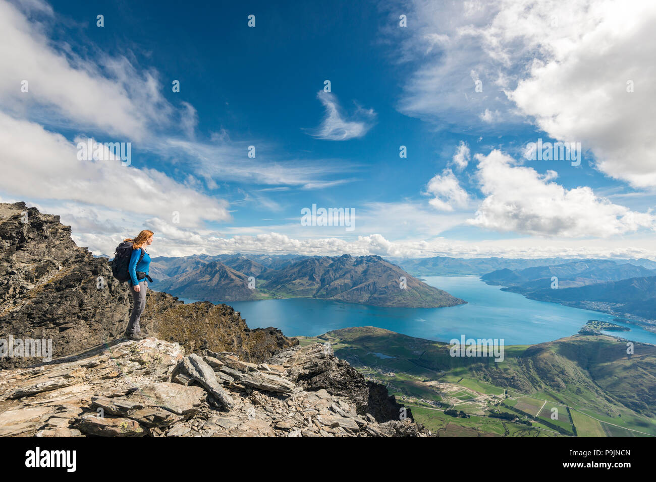 Female hiker stands on rocks in The Remarkables, overlooking Lake Wakatipu, mountains and Queenstown, Queenstown, Otago Stock Photo