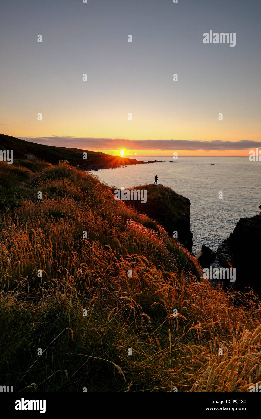 A lone figure standing on the cliff edge watching the sun go down on the South West coast path near St Ives in Cornwall Stock Photo