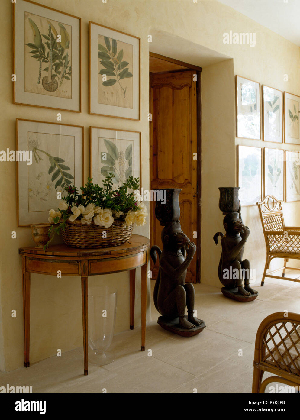 Framed botanical prints above console table with a floral arrangement in a basket in a cream hall with tall oriental statues Stock Photo