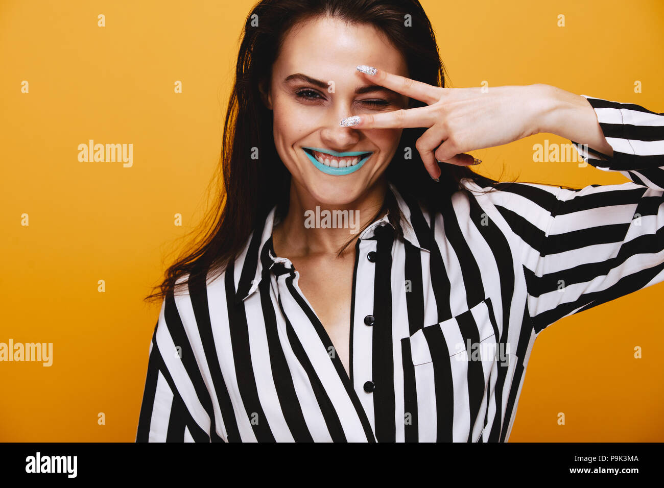 Portrait of smiling female model winking. Woman with artistic makeup wearing a striped dress against orange background. Stock Photo