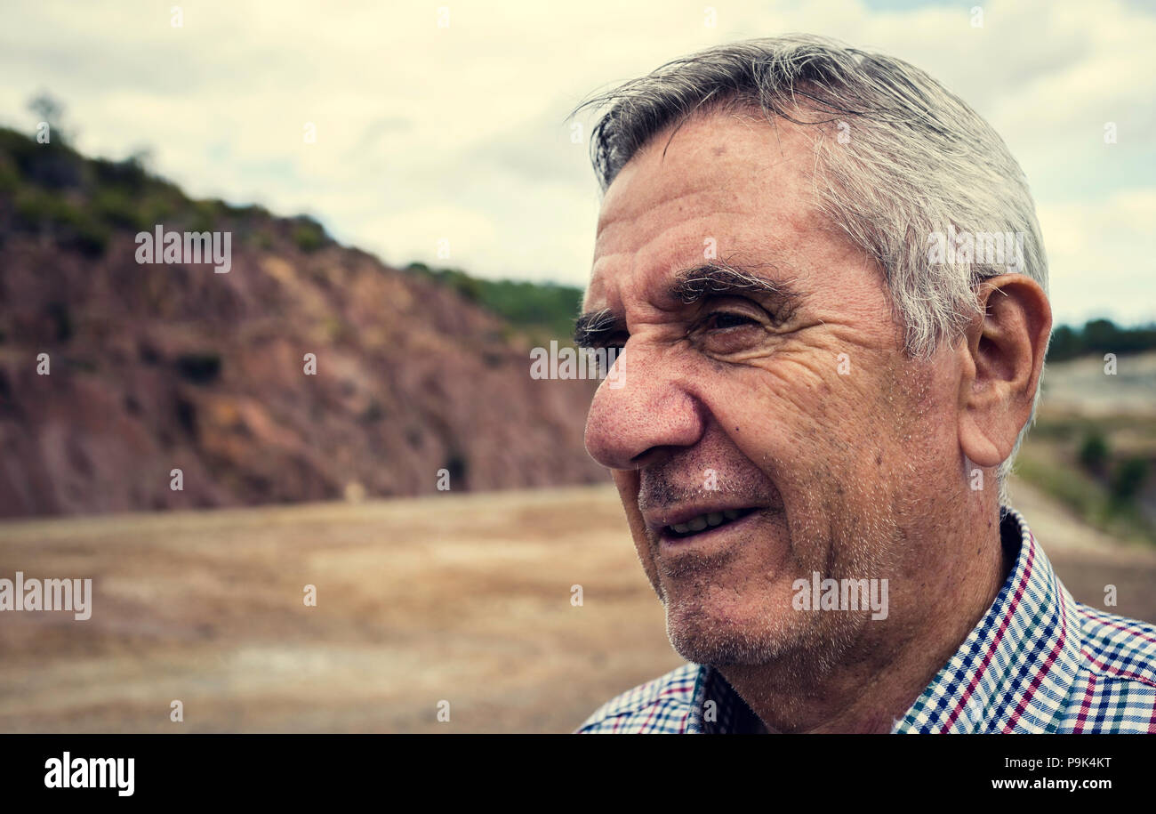 Close-up portrait of an elderly smiling man with white hair and plaid shirt, with Zaranda mines in the background, Spain Stock Photo