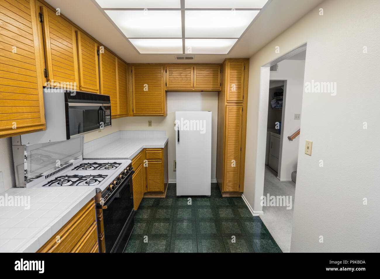 Old 1980s condo kitchen with oak cabinets, tile countertops, gas stove and green flooring. Stock Photo