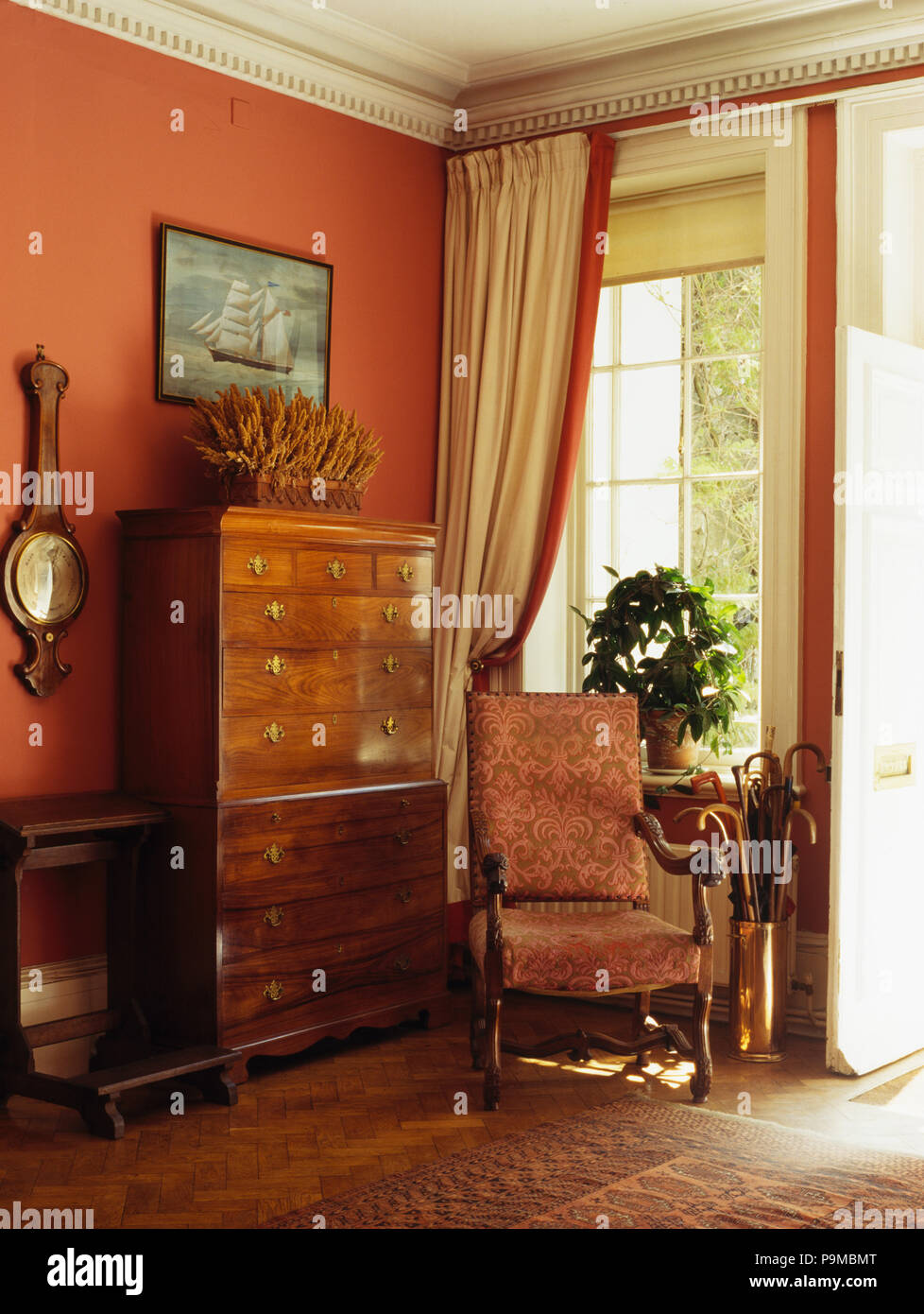 Antique armchair in front of window with cream silk curtain in terracotta country hall with antique chest-of-drawers Stock Photo