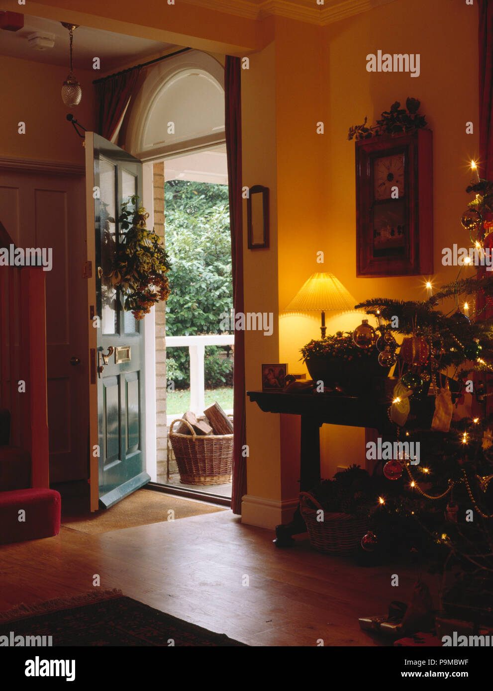 Lighted lamp on table in country hall with Christmas tree and wreath on open front door Stock Photo