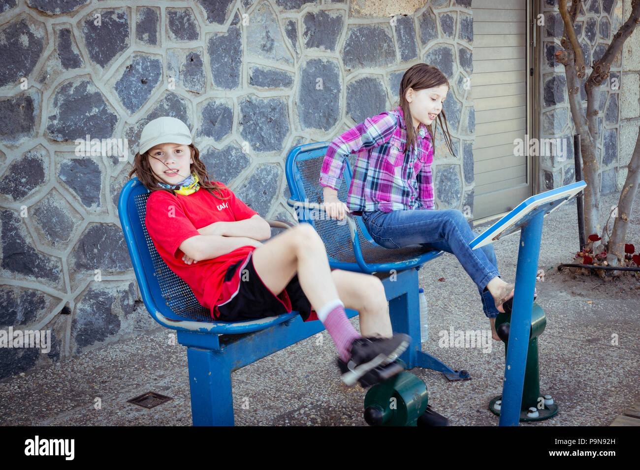 Two caucasian girls children exercising using adult sized outdoor gym equipment, bike pedals with seats, on public street. Stock Photo
