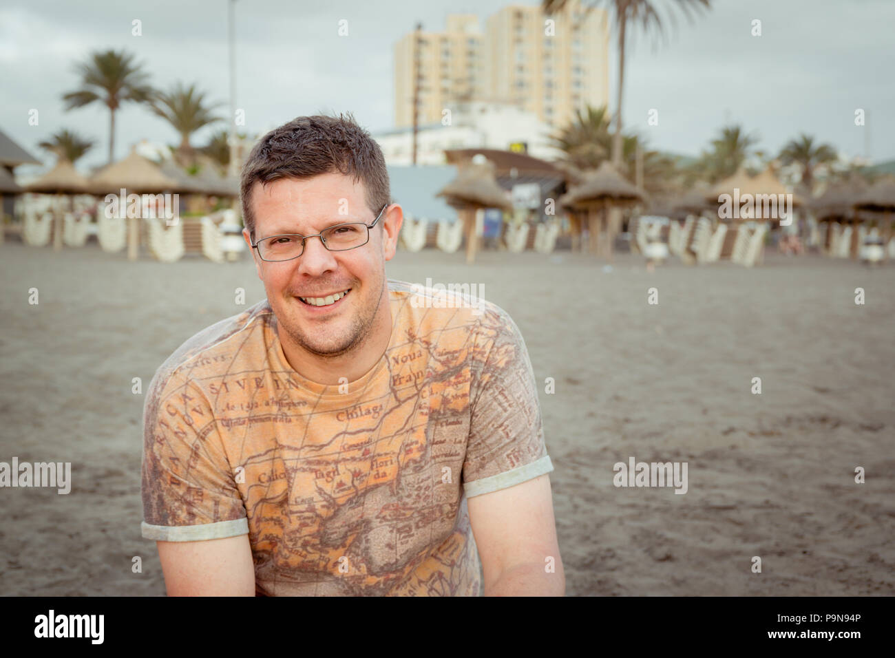 Portrait of a Caucasian man in thirties or forties with brown hair, glasses, stubble wearing a vintage map tshirt on a deserted beach in the evening Stock Photo