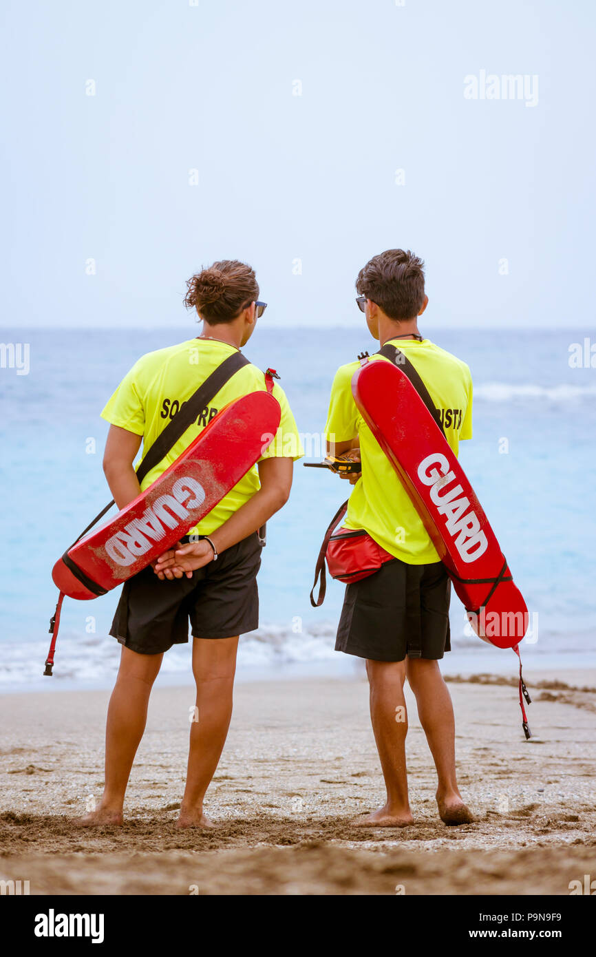 Two adult male Spanish lifeguards patrol a beach on the Tenerife coast (Costa Adeje). With rescue equipment, board, float, walkie talkies. Vertical. Stock Photo