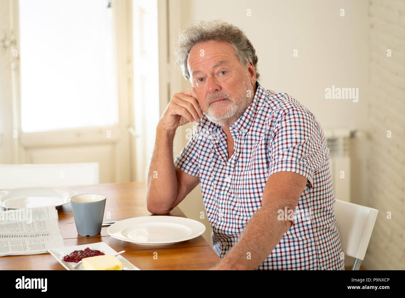Portrait of depressed senior old man with grey hair reading the newspaper while having breakfast and drinking a coffee in old age retirement concept. Stock Photo