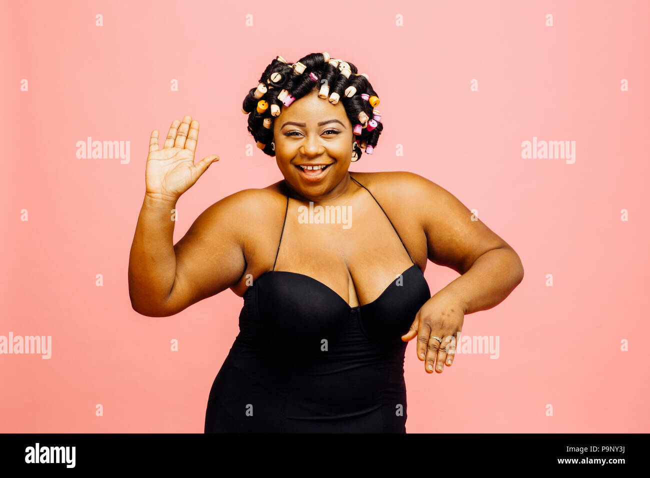 Woman with hair curlers and neglige waiving at camera, isolated on pink background Stock Photo