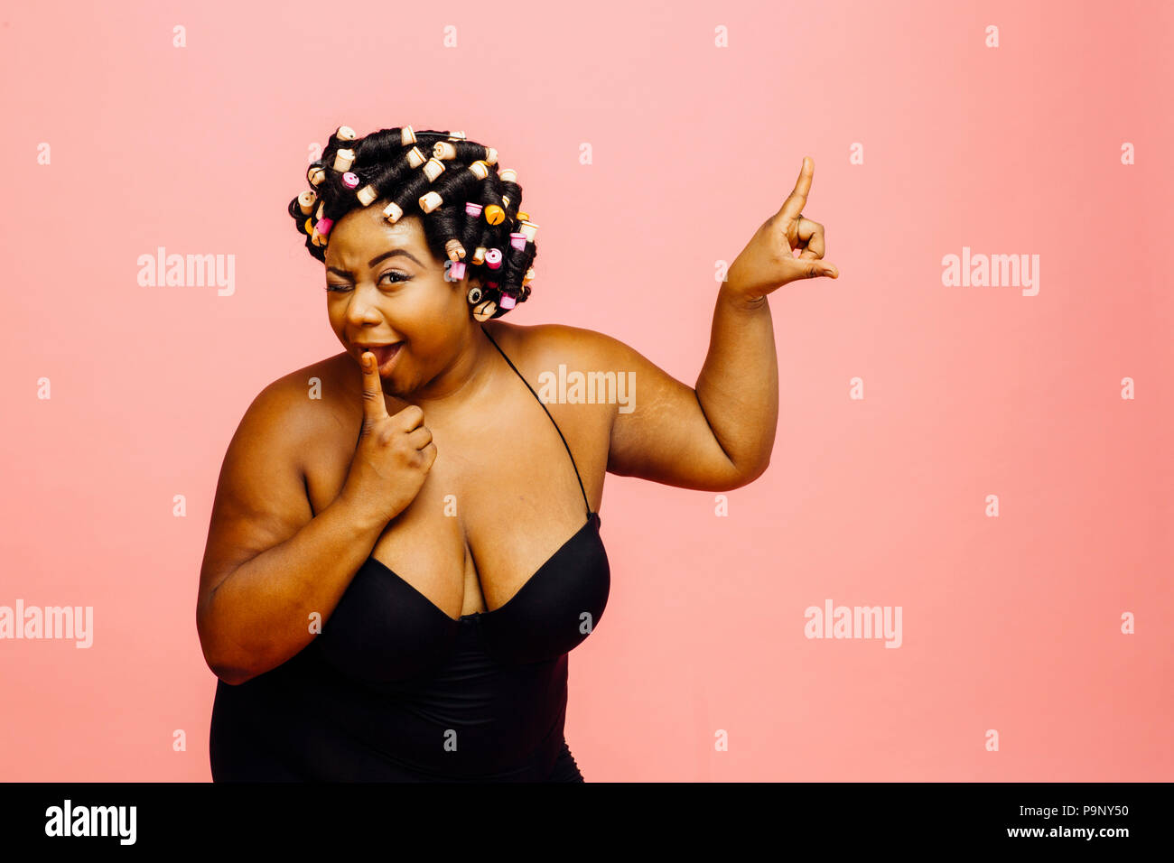 Woman with hair curlers and black camisole, winking and pointing up, isolated on pink background Stock Photo