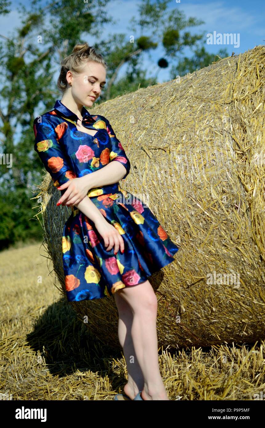 Female model, outdoor photo session, farmland in summer. Girl in colorful dress stands in front of roll of hay. Stock Photo