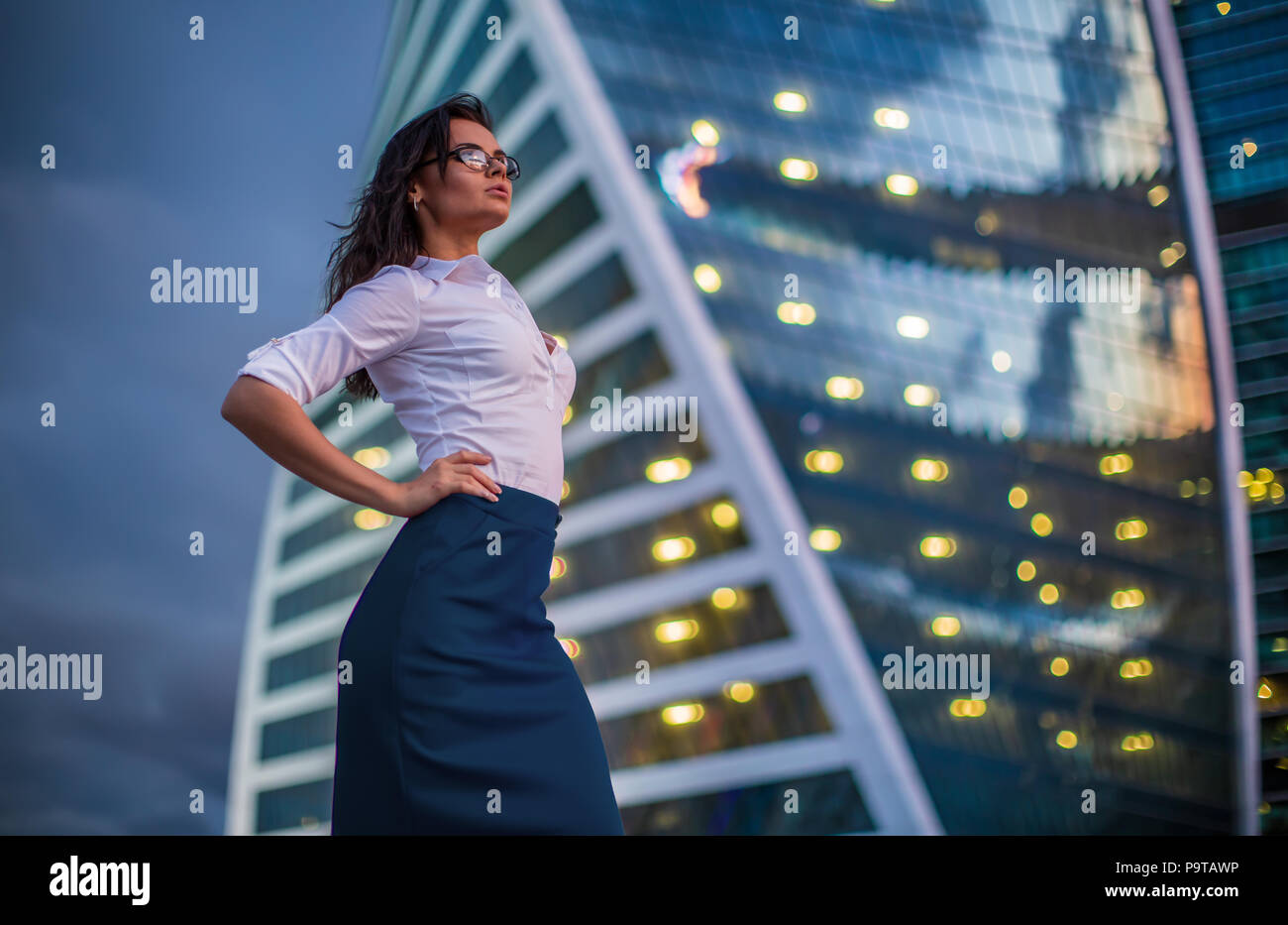 Brunette model wearing glasses, white shirt, grey-blue skirt, standing against business city reflected skyscraper with evening lights flares in window Stock Photo