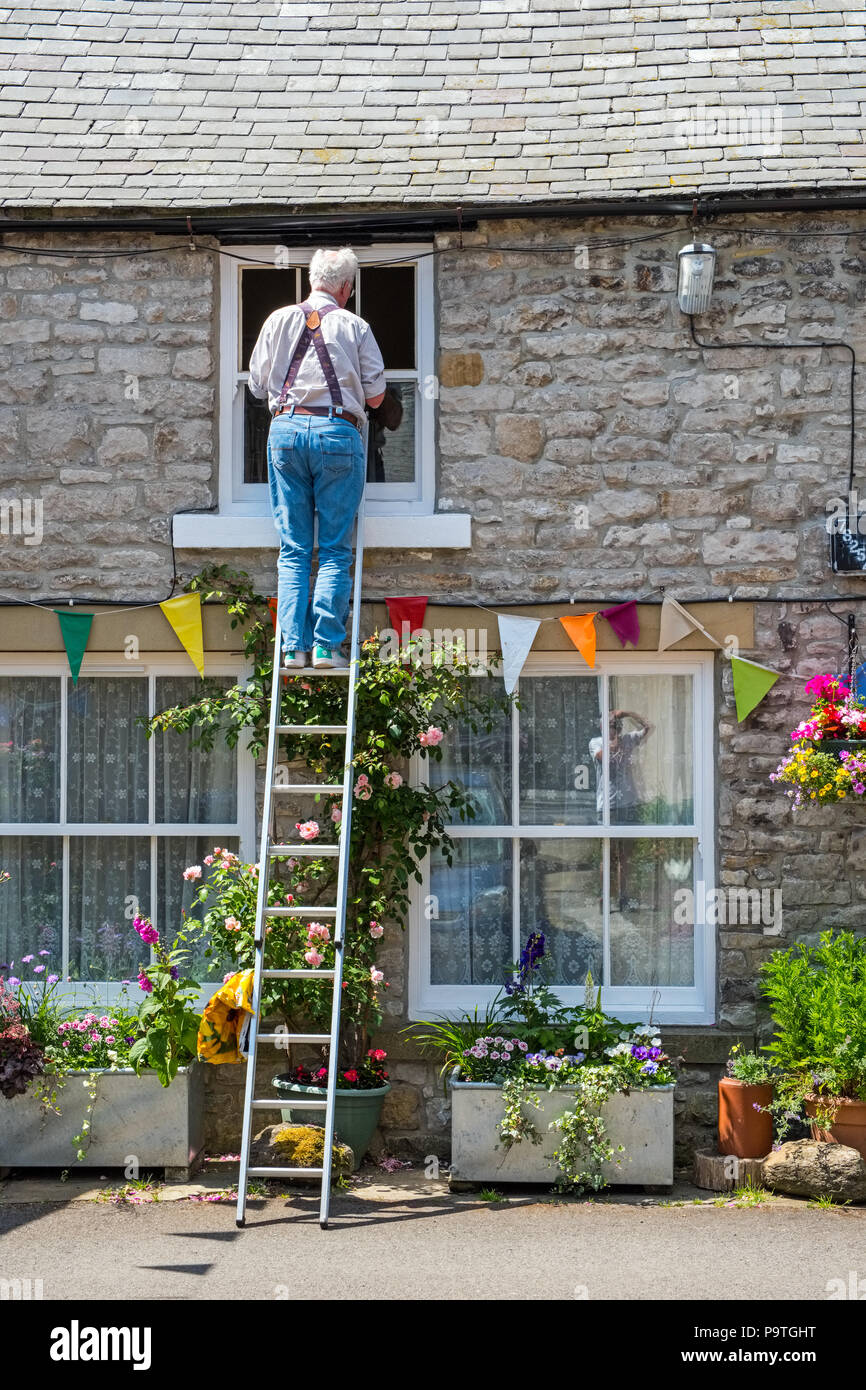 Man up a ladder working on the window of a cottage, Tideswell, Derbyshire Stock Photo