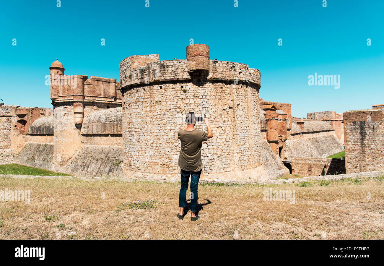 a young caucasian man, seen from behind, taking a picture with his smartphone of the fortress Fort de Salses, built in the 15 century, in Salses-le-Ch Stock Photo