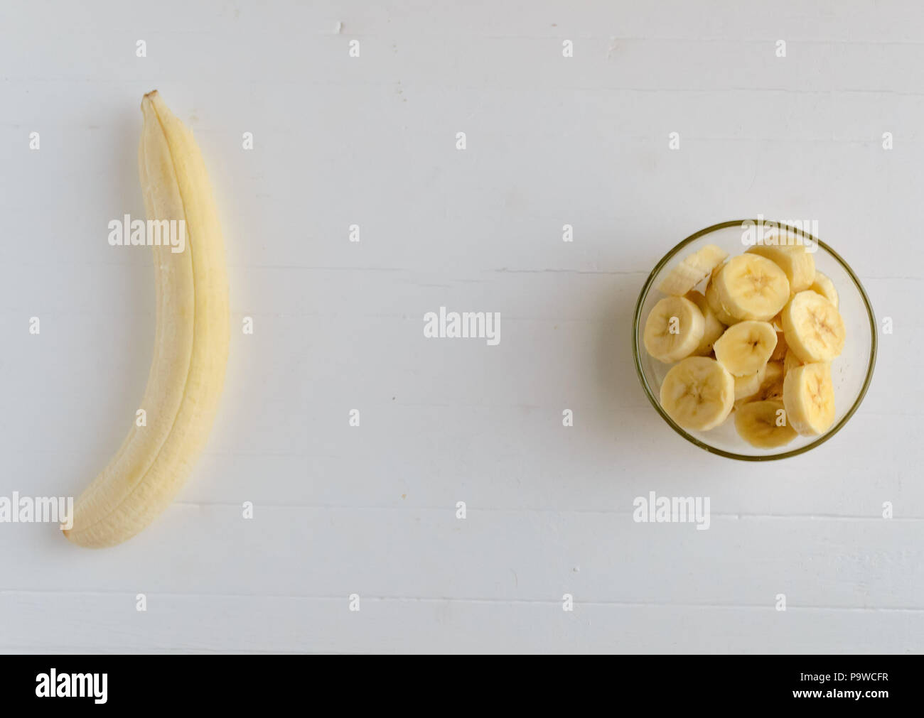 A peeled banana and a fresh ripe organic sliced bananas in a glass bowl on a white wooden background.Copyspace middle. Stock Photo