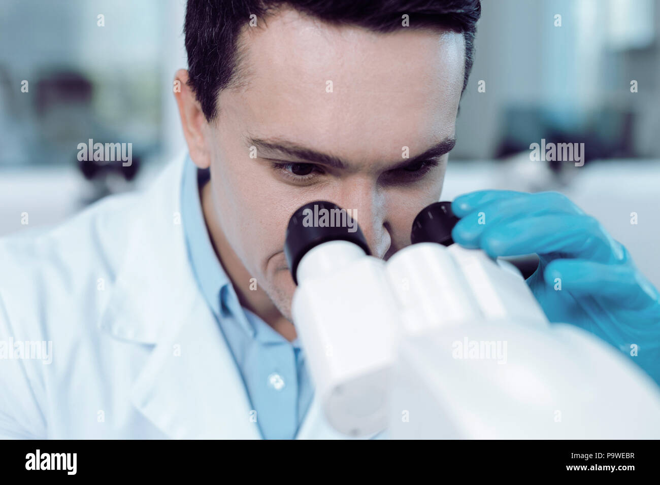 Biological studies. Cheerful positive handsome scientist holding microscope binoculars and looking into them while working on his studies Stock Photo
