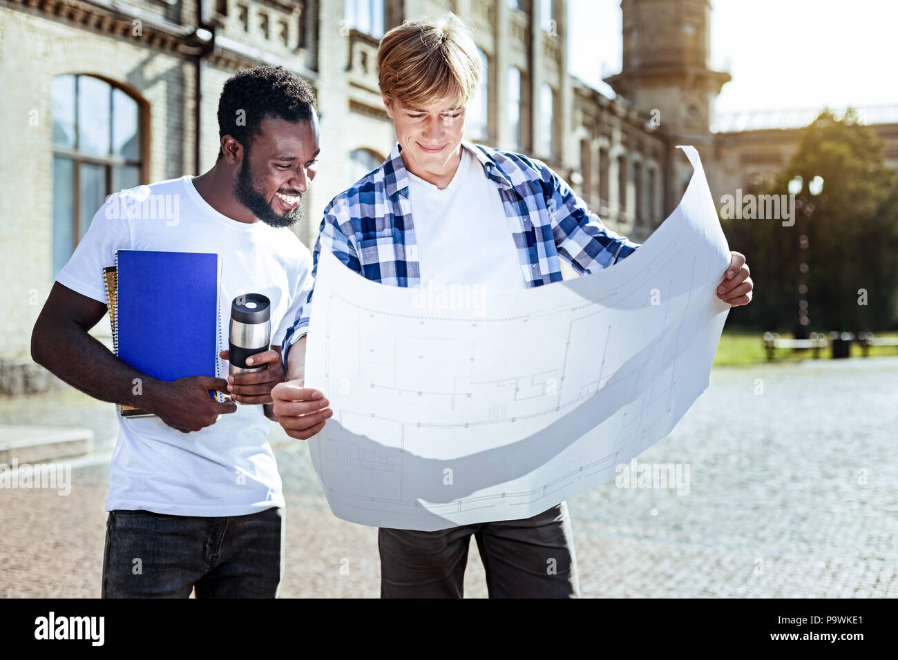 Show it to me. Smiling international student bowing head while looking downwards and standing near his partner Stock Photo