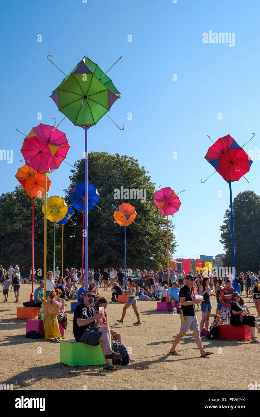 People sit, wander around and enjoy the atmosphere amid a group of stands with brightly coloured umbrellas attached to poles. Latitude Festival. Stock Photo