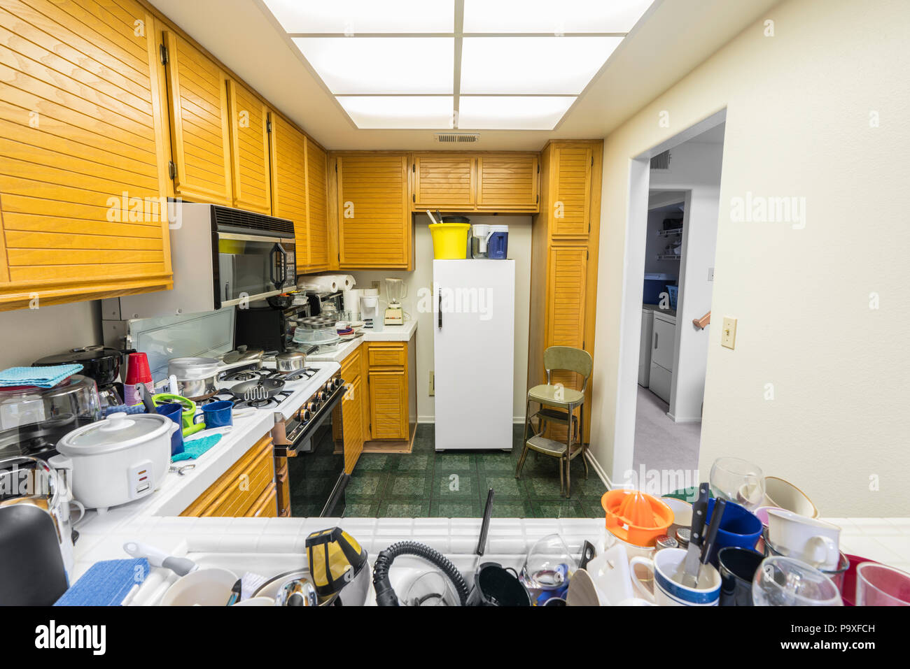 Messy condo kitchen with oak cabinets, tile countertops, gas stove, green flooring and piles of dishes. Stock Photo