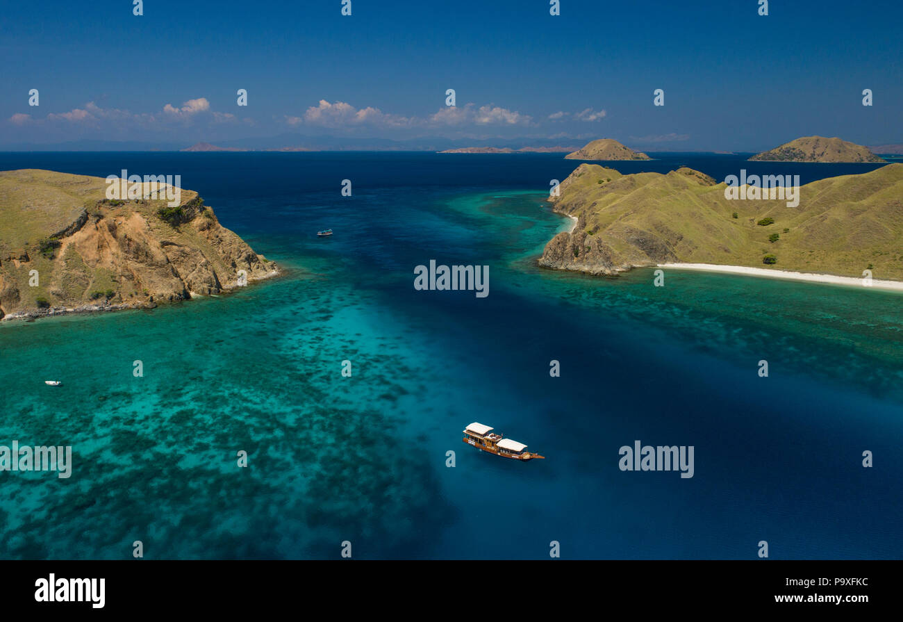 A drone photo of a dive boat over a beautiful turquoise coral reef, with Komodo Island and blue sky in the background. Komodo, Indonesia. Stock Photo