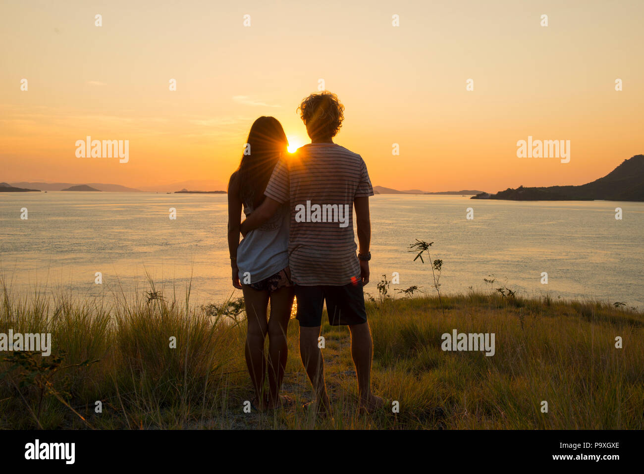 A young tourist couple standing on a headland looking out towards the beautiful sunset over Komodo National Park, Indonesia. Stock Photo