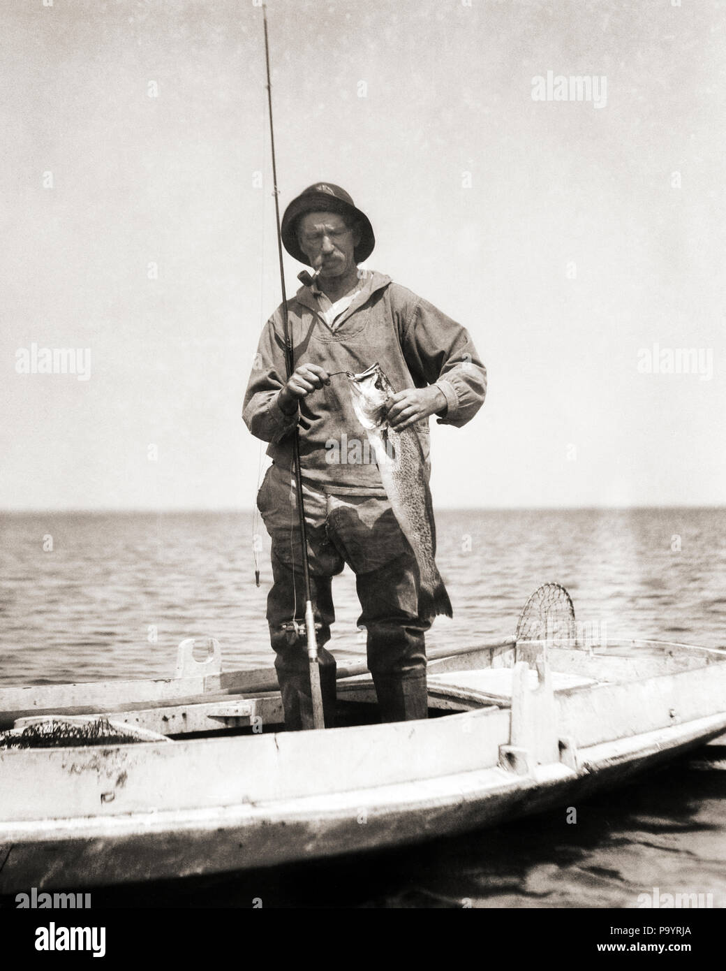 1920s MAN BARNEGAT BAY COMMERCIAL FISHERMAN STANDING IN DUCK BOAT SMOKING PIPE REMOVING FISHHOOK FROM CATCH NEW JERSEY USA - a121 HAR001 HARS SUCCESS REMOVING NJ OCCUPATIONS DUCK BOAT FISHING ROD NEW JERSEY BARNEGAT BAY BLACK AND WHITE CAUCASIAN ETHNICITY HAR001 OLD FASHIONED Stock Photo
