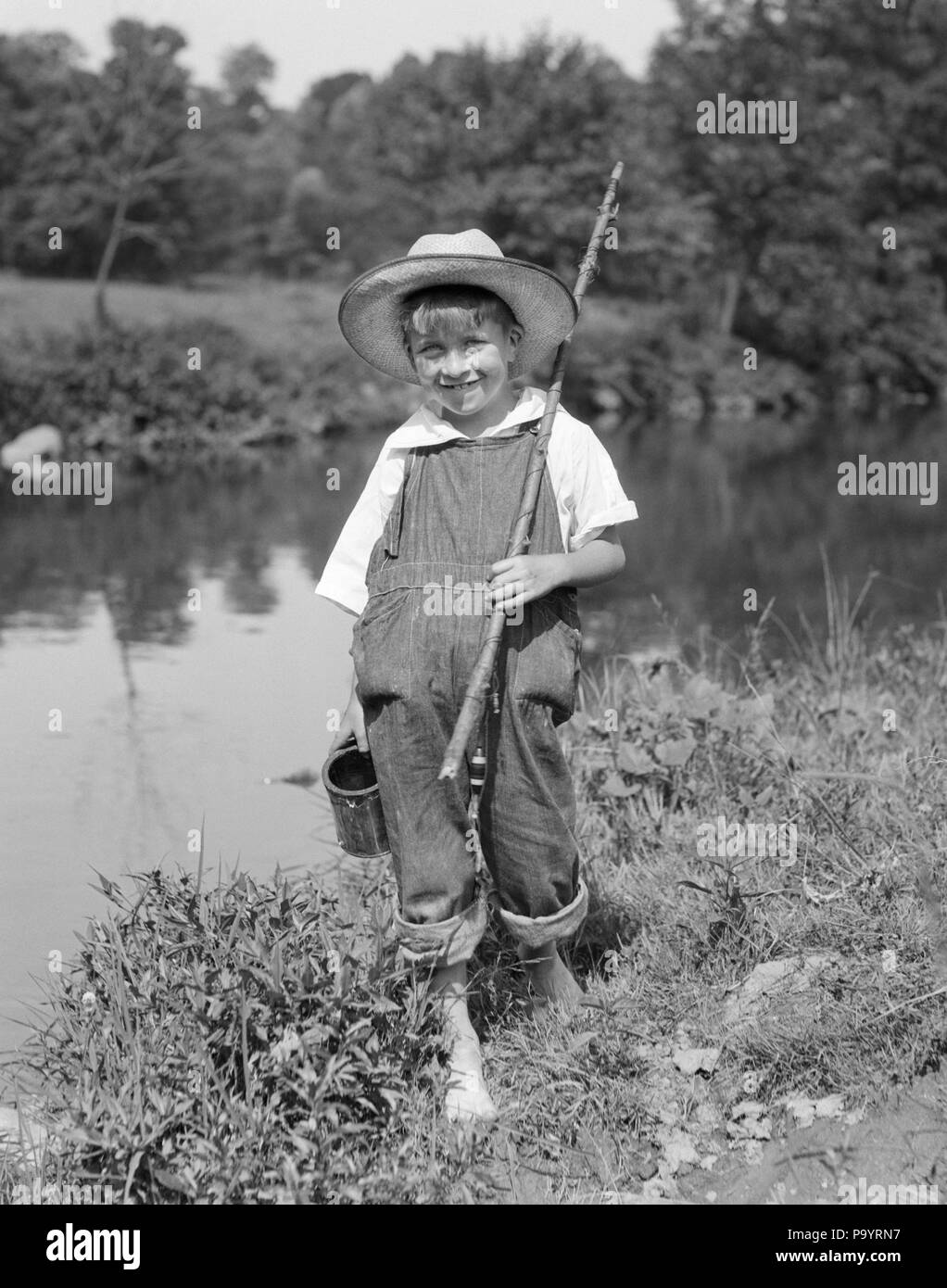 1920s 1930s BAREFOOT BOY CARRYING STICK FISHING ROD CAN OF BAIT WORMS  WEARING STRAW HAT BIB OVERALLS LOOKING AT CAMERA SMILING - a4795 HAR001  HARS COPY SPACE FULL-LENGTH OVERALLS STREAM MALES ROD