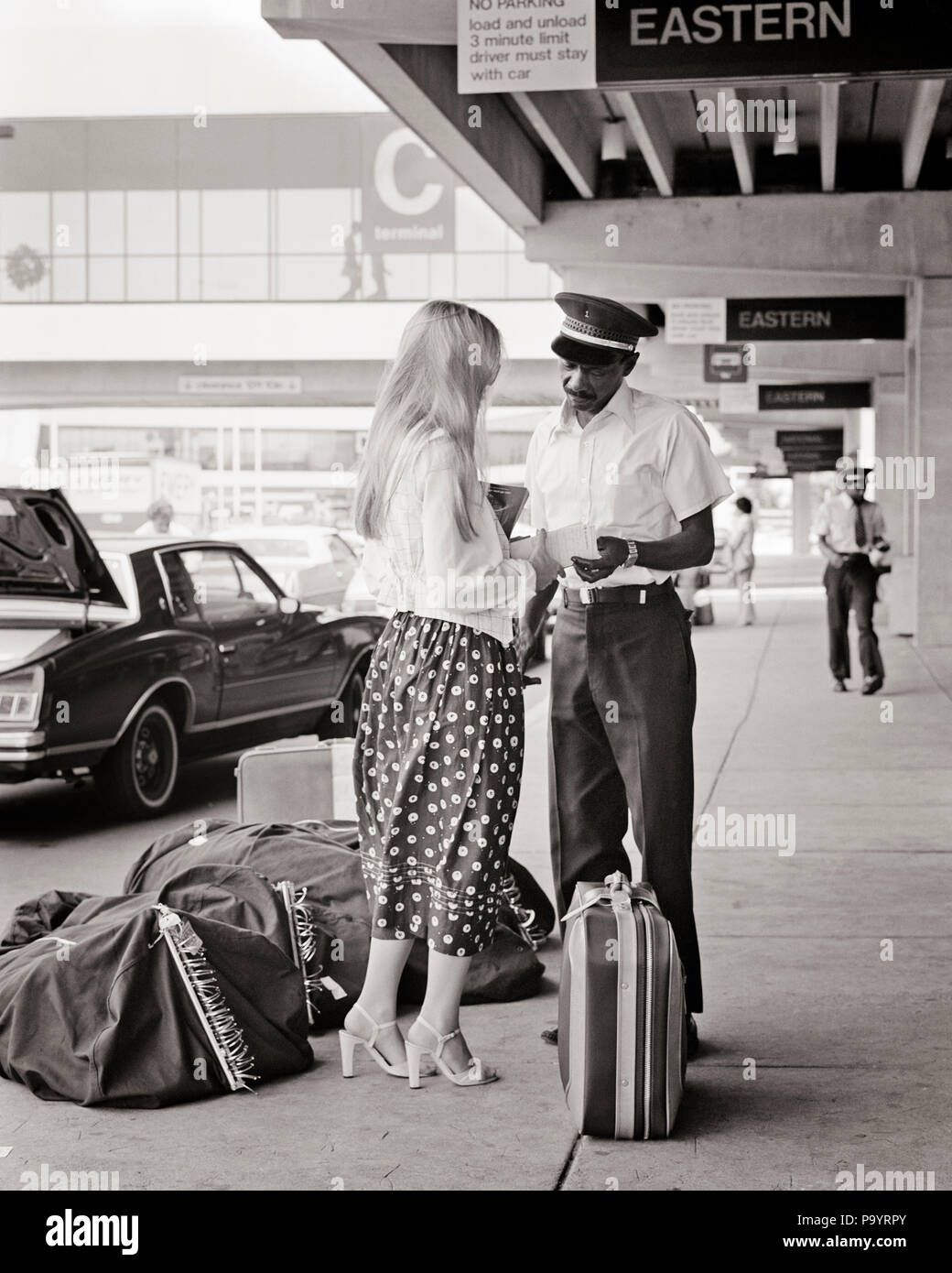 1970s BLONDE WOMAN CHECKING BAGS CURBSIDE WITH  AFRICAN AMERICAN AIRPORT SKYCAP - a8596 HAR001 HARS INFORMATION CHECKING LIFESTYLE AIRLINE FEMALES JOBS COPY SPACE FULL-LENGTH LADIES PERSONS MALES TRANSPORTATION B&W OCCUPATION ADVENTURE STYLES CUSTOMER SERVICE AFRICAN-AMERICANS AFRICAN-AMERICAN BLACK ETHNICITY LABOR EMPLOYMENT OCCUPATIONS TRAVELING CONNECTION STYLISH CURBSIDE EMPLOYEE FASHIONS MID-ADULT MID-ADULT MAN TRAVELER YOUNG ADULT WOMAN BAGGAGE BLACK AND WHITE CAUCASIAN ETHNICITY HAR001 LABORING OLD FASHIONED AFRICAN AMERICANS Stock Photo