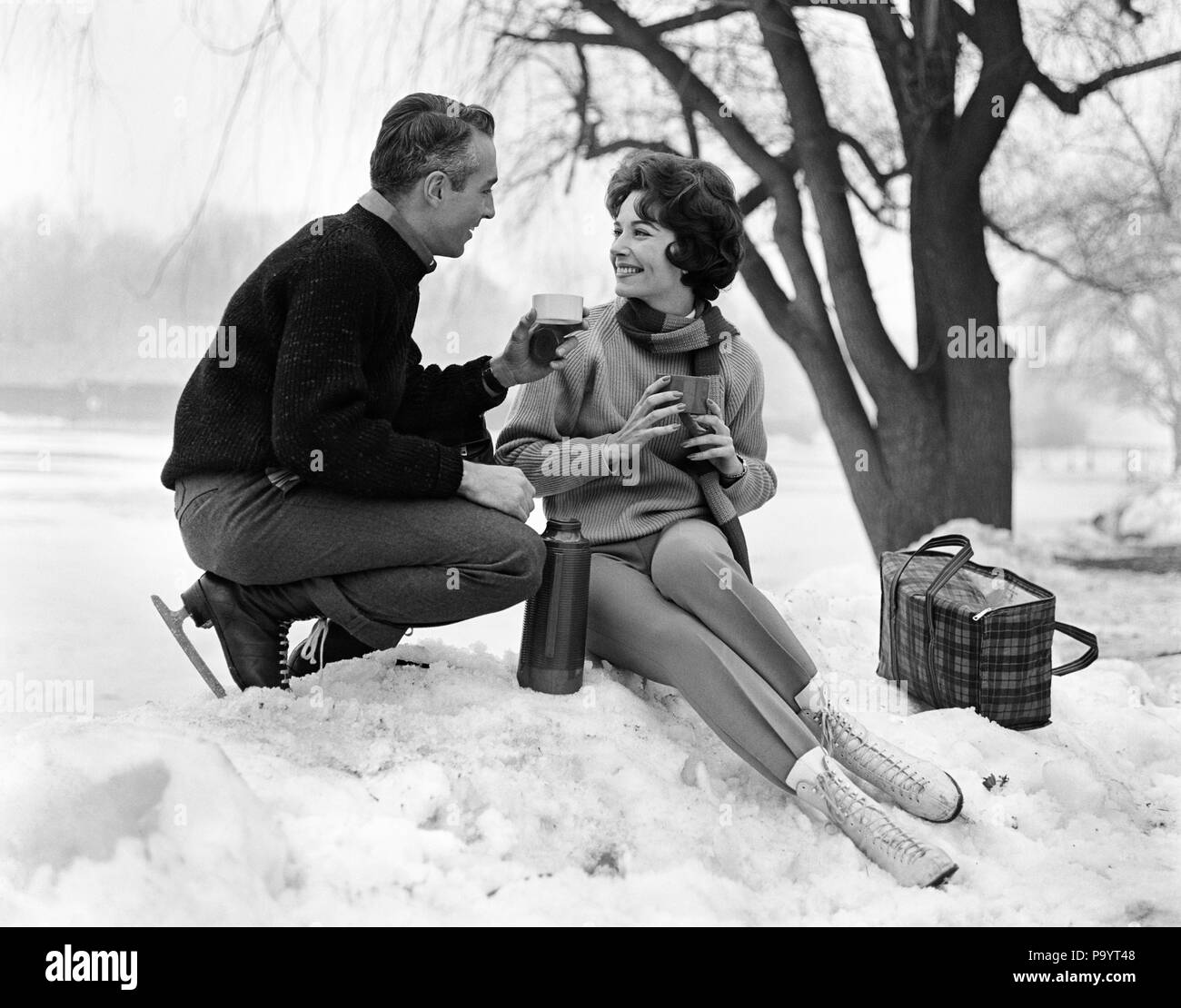 1960s SMILING COUPLE IN SNOW WEARING ICE SKATES DRINKING HOT BEVERAGE FROM THERMOS - bw00113 CAM001 HARS FRIENDSHIP HALF-LENGTH LADIES PERSONS MALES B&W WINTERTIME DATING HAPPINESS CHEERFUL EXCITEMENT RECREATION CAM001 SMILES JOYFUL STYLISH MID-ADULT MID-ADULT MAN MID-ADULT WOMAN TOGETHERNESS WIVES BLACK AND WHITE CAUCASIAN ETHNICITY OLD FASHIONED Stock Photo