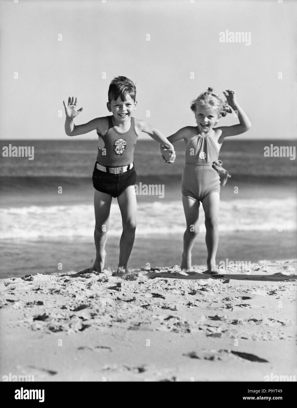 1930s TWO KIDS BOY GIRL HOLDING HANDS RUNNING ON SANDY BEACH - bx001393 CAM001 HARS FRIENDSHIP FULL-LENGTH PERSONS MALES SIBLINGS SURF SISTERS B&W SUMMERTIME EYE CONTACT SHORE HAPPINESS EXCITEMENT RECREATION CAM001 HOLDING HANDS SIBLING SANDY BATHING SUIT SWIM WEAR GROWTH JUVENILES SEASHORE TOGETHERNESS BLACK AND WHITE CAUCASIAN ETHNICITY OLD FASHIONED Stock Photo