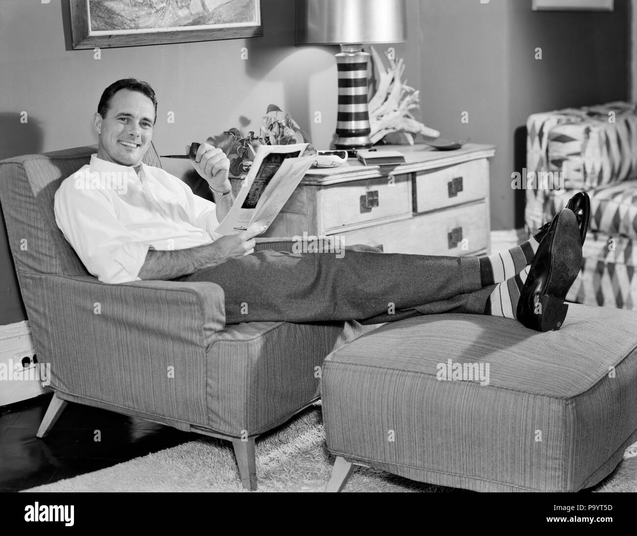 1950s RELAXED MAN SITTING IN EASY CHAIR FEET UP SMOKING PIPE READING MAGAZINE LOOKING AT CAMERA - bx016518 CAM001 HARS CAM001 SMILES EASY JOYFUL OTTOMAN STYLISH MID-ADULT MID-ADULT MAN RELAXATION BLACK AND WHITE CAUCASIAN ETHNICITY OLD FASHIONED Stock Photo
