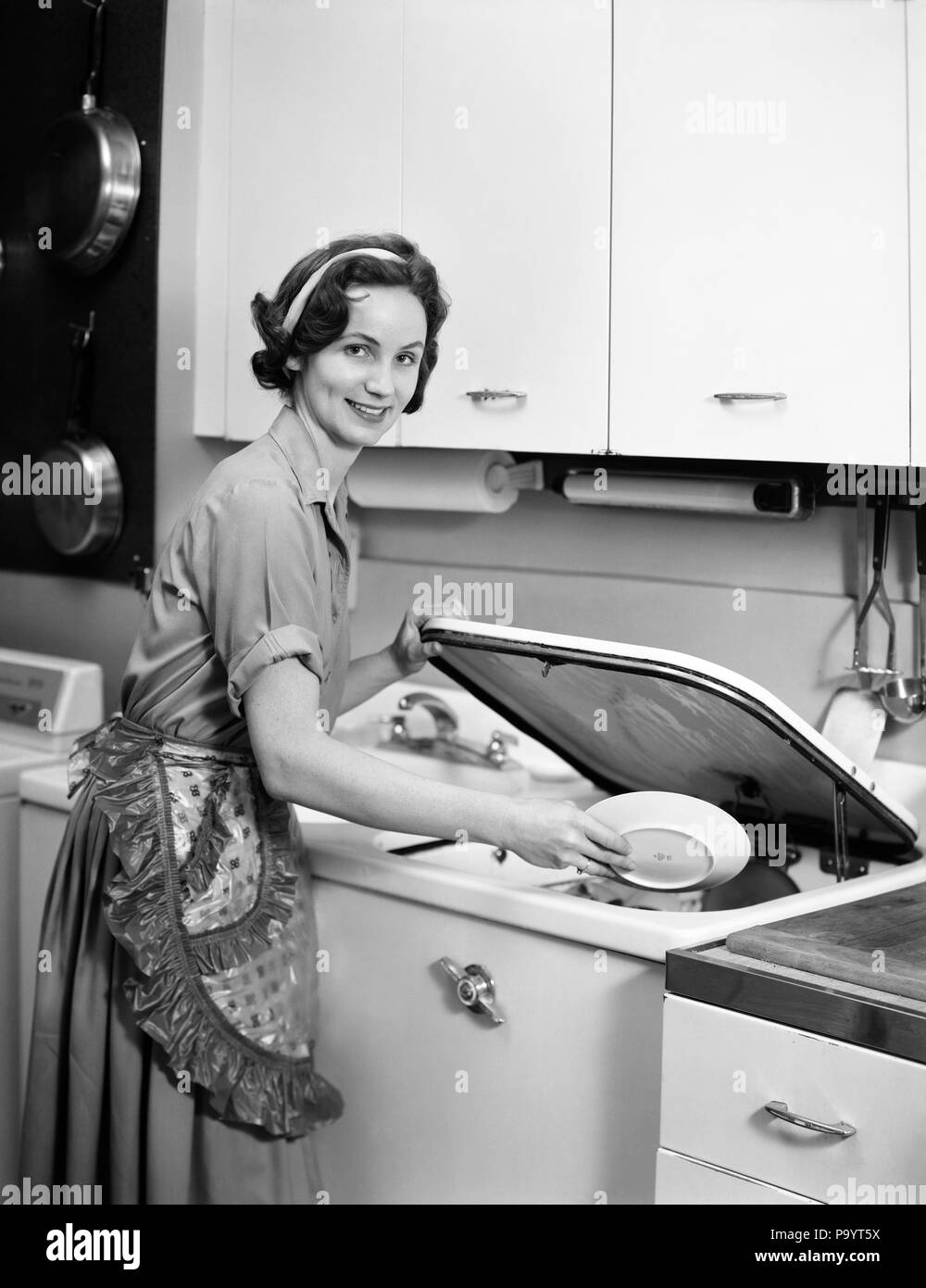 1950s 1960s SMILING HOUSEWIFE LOADING DISHWASHER LOOKING AT CAMERA - bx018415 CAM001 HARS CAM001 HOUSEWIVES SMILES JOYFUL MID-ADULT MID-ADULT WOMAN YOUNG ADULT WOMAN BLACK AND WHITE CAUCASIAN ETHNICITY OLD FASHIONED Stock Photo