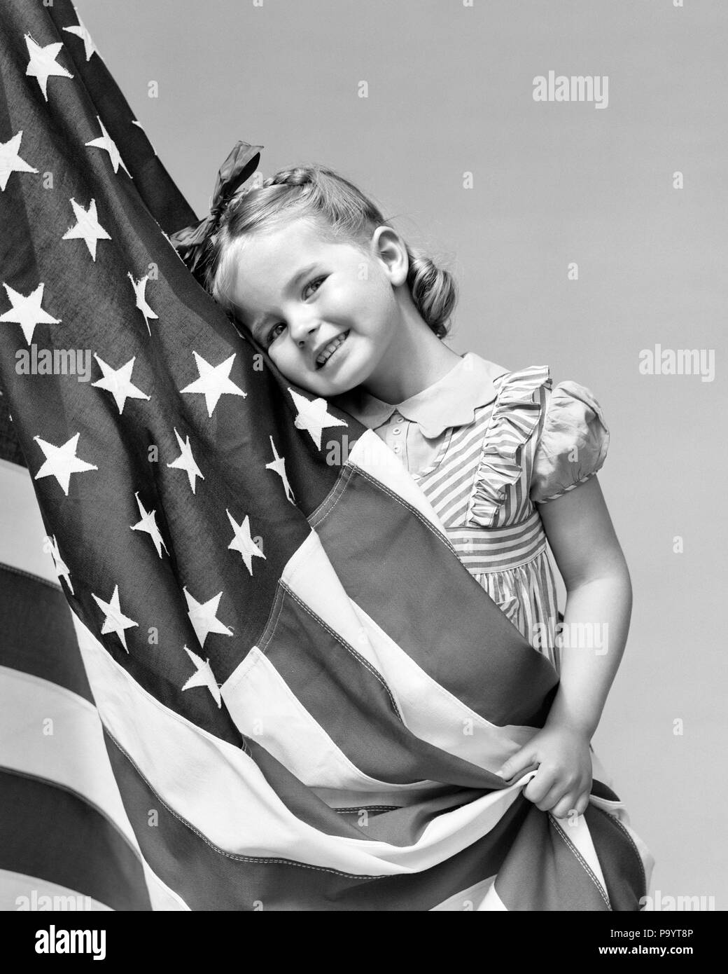 1940s LITTLE GIRL HUGGING THE AMERICAN FLAG SMILING LOOKING AT CAMERA - h1281 HAR001 HARS PATRIOTIC STARS AND STRIPES SUPPORT PATRIOTISM AMERICAN FLAG BLACK AND WHITE CAUCASIAN ETHNICITY HAR001 OLD FASHIONED Stock Photo