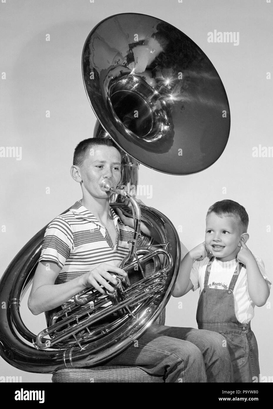 1950s TEEN BOY IN STRIPE SHIRT JEANS PLAYING TUBA SMALLER BROTHER IN BIB OVERALLS SMILING INDEX FINGERS STUCK IN BOTH EARS  - m11569 TAY001 HARS JEANS EXPRESSION OLD TIME NOSTALGIA BROTHER OLD FASHION 1 JUVENILE FACIAL EARS COMMUNICATION INSTRUMENTS COMIC SAFETY FINGERS NOISE FAMILIES LIFESTYLE HORN SOUND MUSICIAN BROTHERS HOME LIFE COMMUNICATING STRIPE HALF-LENGTH PERSONS OVERALLS MALES RISK TEENAGE BOY ENTERTAINMENT SIBLINGS EXPRESSIONS B&W HORNS MUSICIANS OVERSIZED PERFORMING ARTS INDEX WEIRD BOTH LEISURE MANUAL PROTECTION STUCK LOUD OVERSIZE POWERFUL ZANY UNCONVENTIONAL SIBLING SMILES Stock Photo