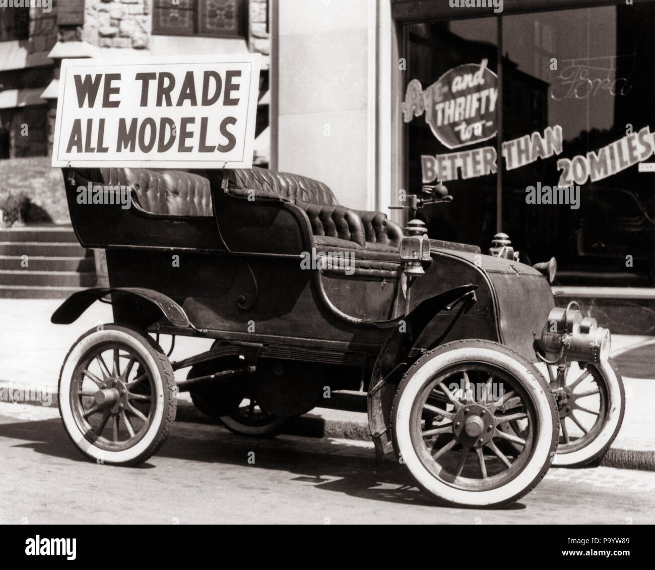 1930s ANTIQUE CAR WITH WE TRADE ALL MODELS ADVERTISING SIGN PARKED ON STREET IN FRONT OF CAR DEALERSHIP - m1583 HAR001 HARS BLACK AND WHITE HAR001 OLD FASHIONED Stock Photo