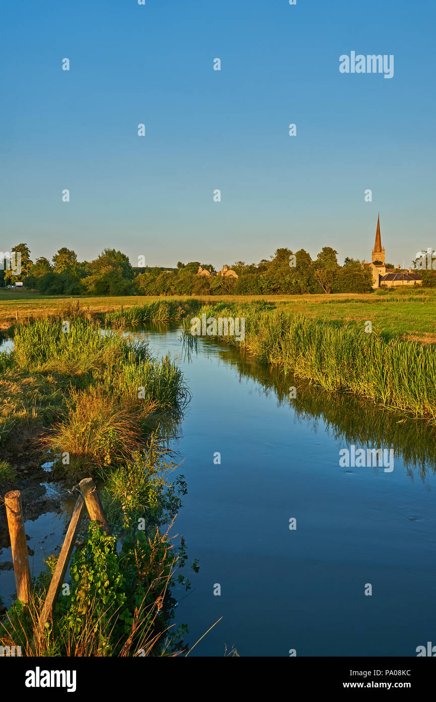 The River Windrush winding through water meadows towards the Cotswold town of Burford, with the church spire of St John the Baptist in the distance. Stock Photo