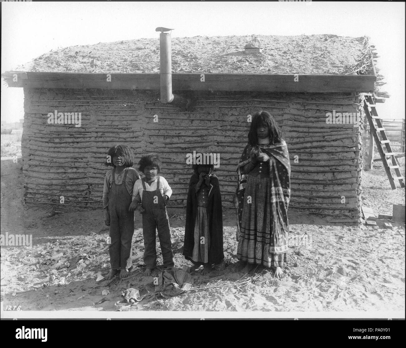 . English: Four Mojave Indian children standing in front of a small dwelling with a thatched roof, near Yuma, ca.1900 Photograph of 4 Mojave Indian children standing in front of a small dwelling with a thatched roof, near Yuma, ca.1900. The 2 little boys are wearing overalls, the 2 little girls are both wearing blankets or large shawls reaching to the ground over their shoulders, and long dresses. A stove pipe sticks out of the wall of the dwelling behind them. A ladder is visible behind the dwelling at right.  Call number: CHS-1278 Photographer: Pierce, C.C. (Charles C.), 1861-1946 Filename:  Stock Photo