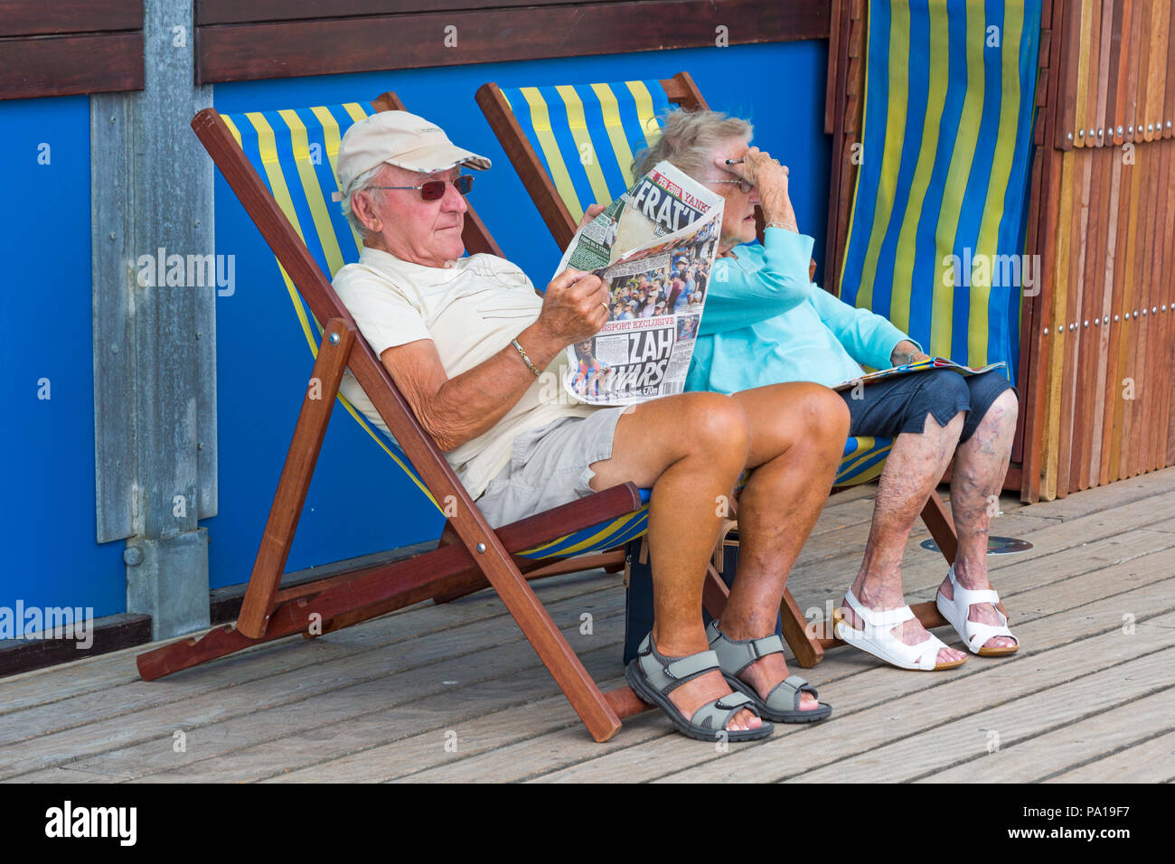 Bournemouth, Dorset, UK. 20th July 2018. UK weather: hot and humid with hazy sunshine at Bournemouth, as sunseekers head to the seaside at Bournemouth beaches to enjoy the fine weather. Senior couple sitting in deckchairs on Bournemouth pier, the man reading The Sun newspaper.  Credit: Carolyn Jenkins/Alamy Live Stock Photo