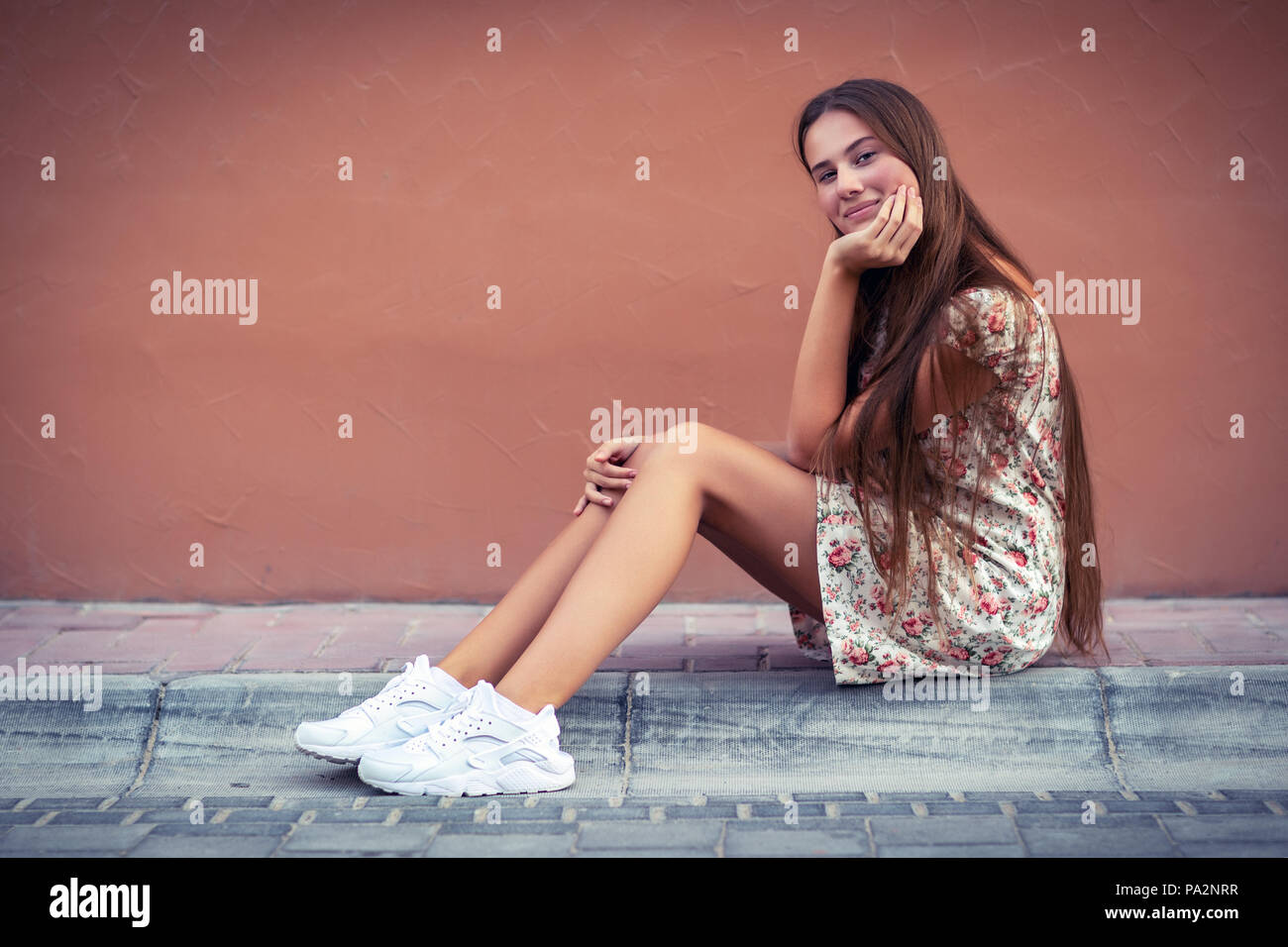 Pretty girl on the street, nice female with long natural hair sitting on blank monophonic wall background, authentic genuine beauty of young people Stock Photo