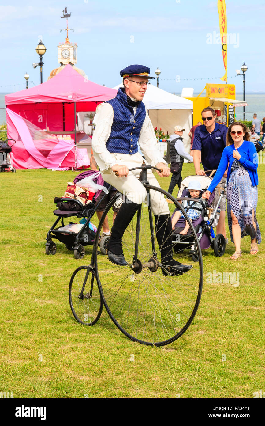 Man dressed up in Victorian costume as a Charles Dickens character riding around park on a penny farthing bike, watched by young family. Stock Photo