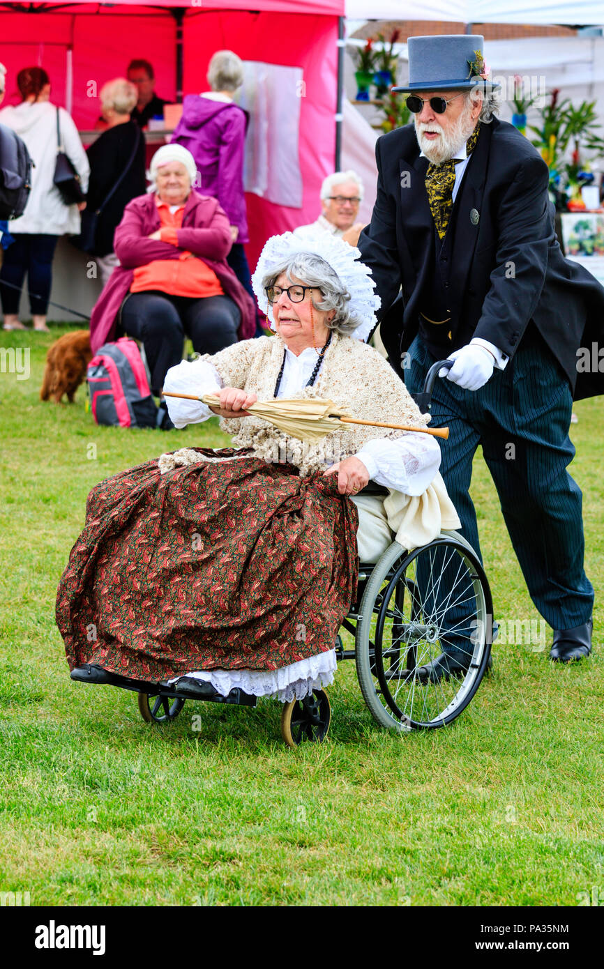 Senior woman in wheelchair being pushed by gentleman in black suit. Wearing Victorian 19th century costume, clothing. She holds parasol, looks angry. Stock Photo