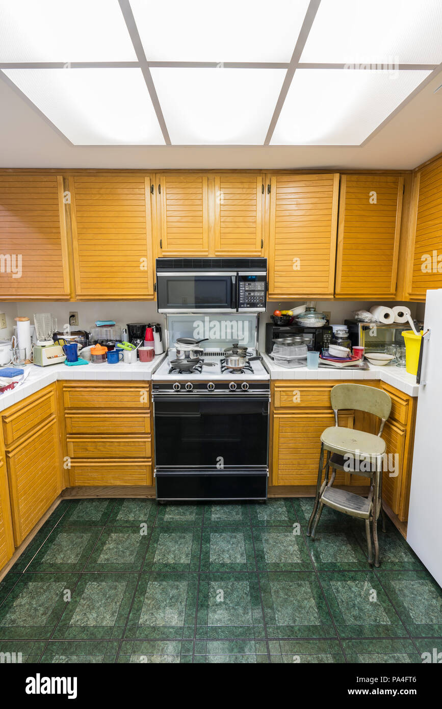 Messy condo kitchen vertical view with oak cabinets, tile countertops, gas stove, green flooring and piles of dishes. Stock Photo