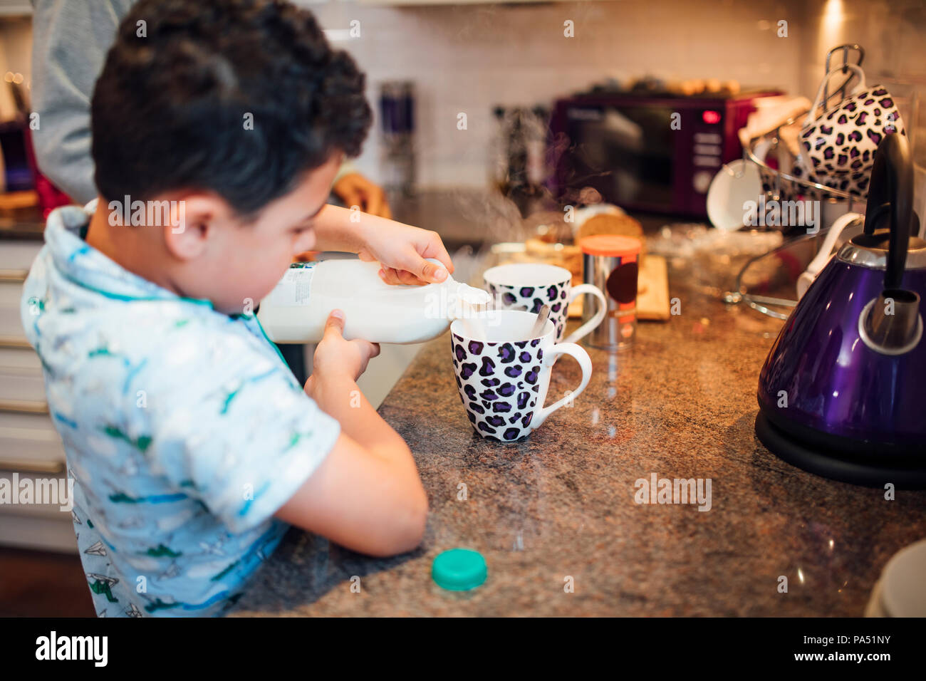 Little boy is pouring milk in to a mug while making tea with his father at home. Stock Photo