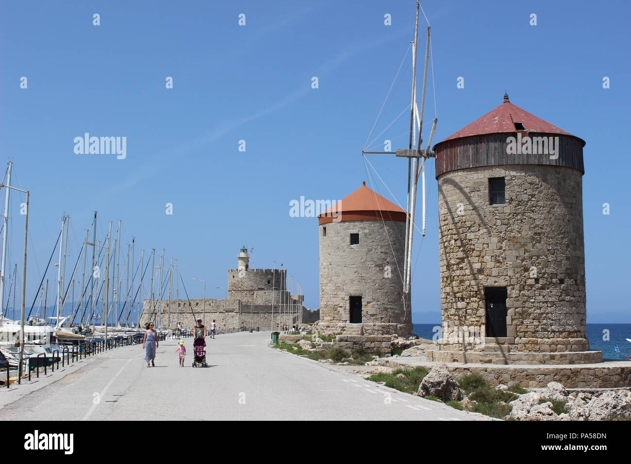 Old windmills at the center of the Rhodes, Faliraki. A tourist family enjoys beautiful shinny day by sightseeing along the walking way. Stock Photo