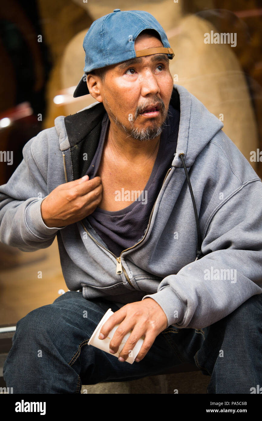 Native American man sitting on sidewalk bench wearing light blue ball cap grey hooded sweatshirt looking to upper right scratching his chest. Stock Photo