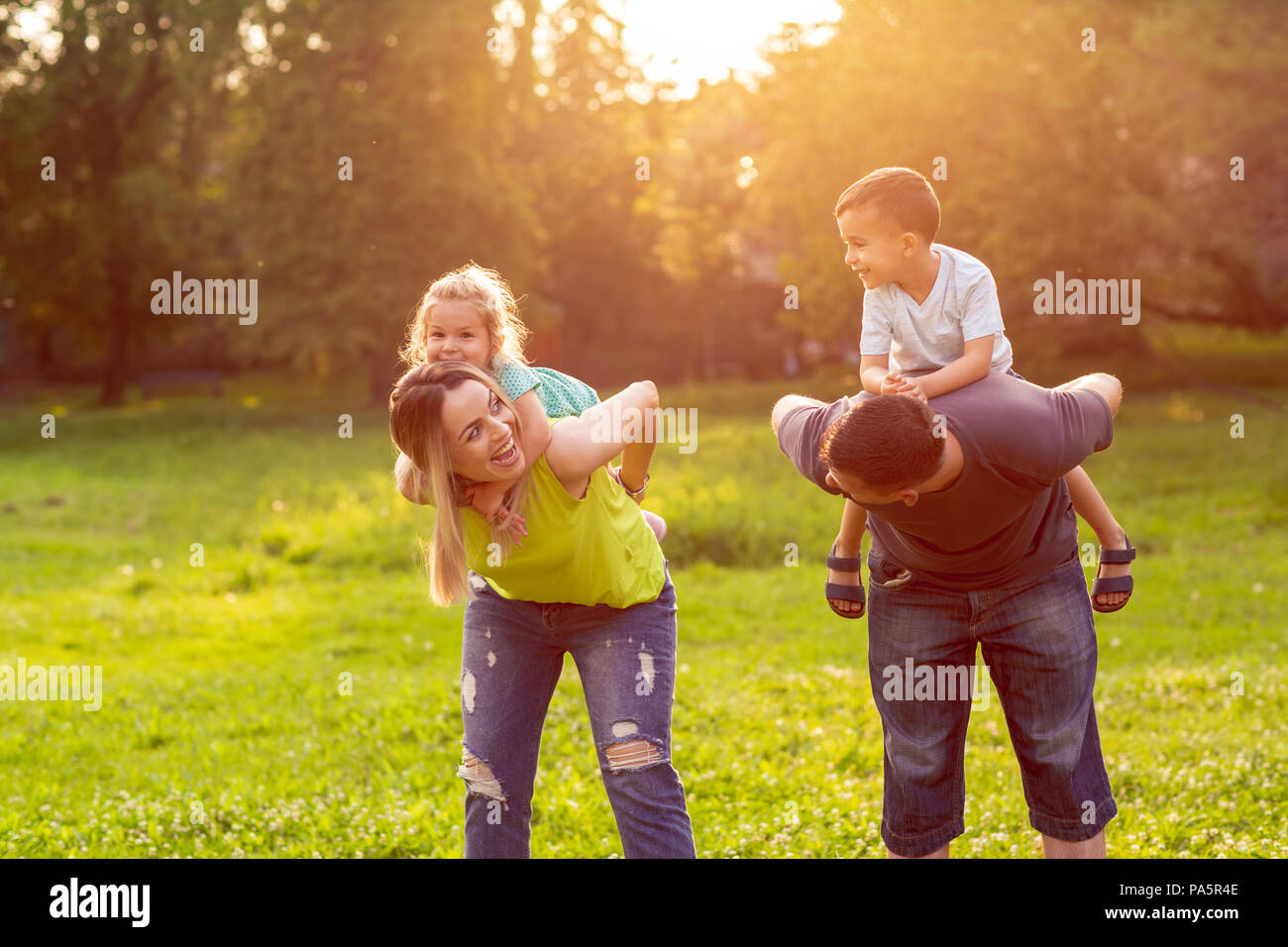 family, happiness, childhood and people concept - Happy parents giving piggyback ride to children Stock Photo