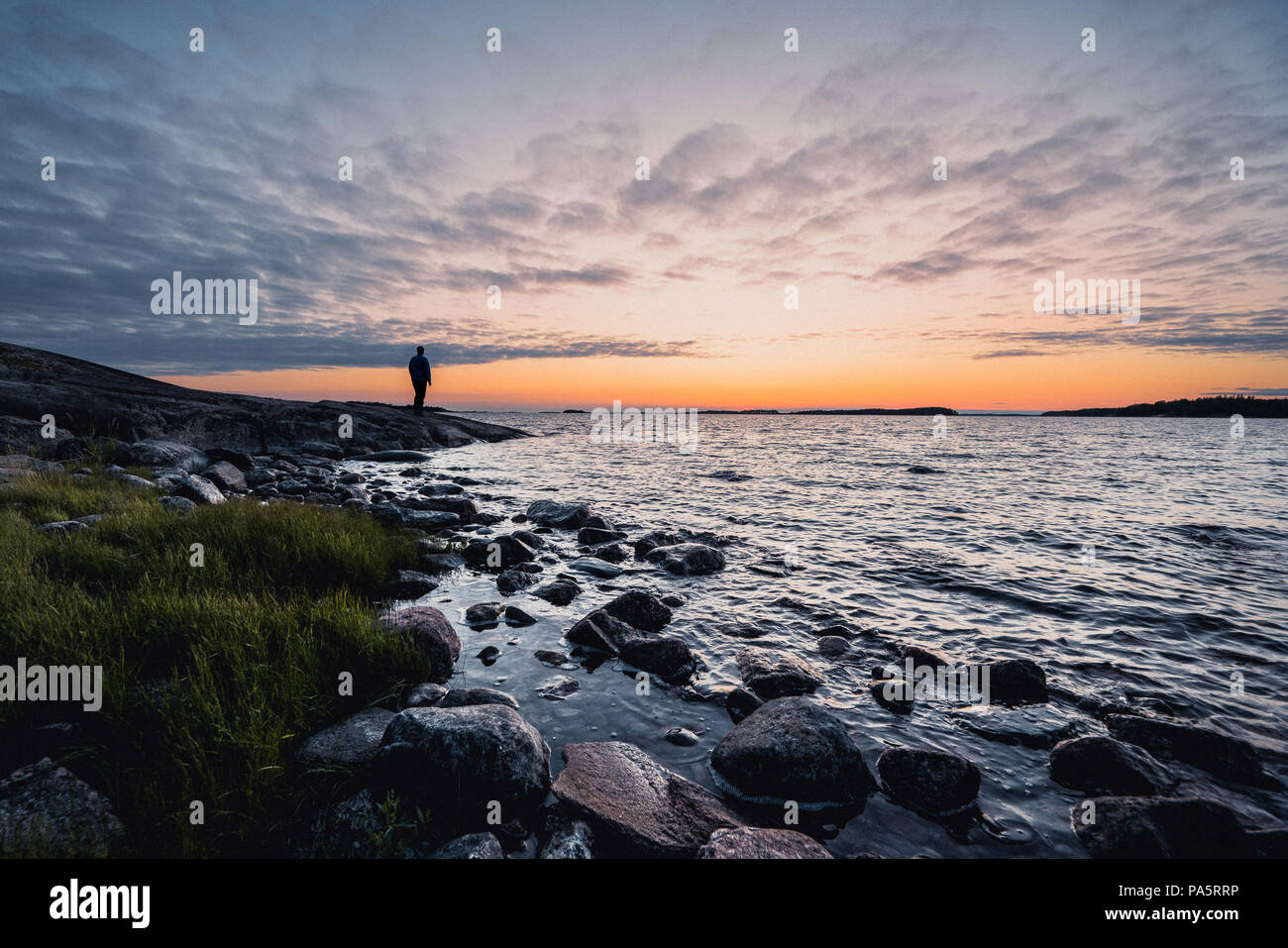 Man standing alone on a rock by a windy sea and looking at the horizon and sunset Stock Photo